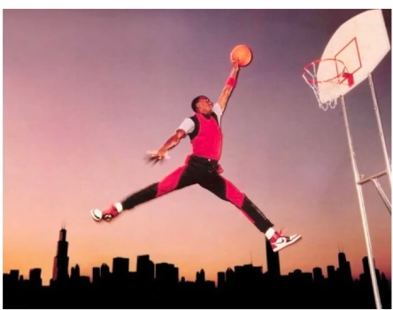 A basketball player in a red and black uniform leaps high in the air against a sunset sky, legs spread in a split, holding a basketball overhead toward the hoop. The city skyline, including the Sears Tower, is silhouetted in the background.