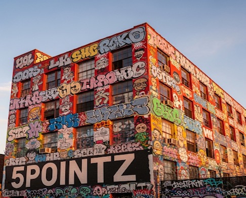 An AI produced image of a large industrial building covered in colorful graffiti murals and tags. The walls feature vibrant artwork, lettering, and characters in bold styles, with windows partially painted over. Prominently displayed across the lower section of the building is the word “5POINTZ” in large white block letters outlined in black..